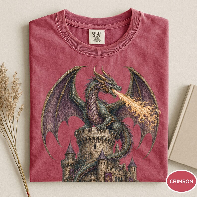 May include: A crimson short-sleeved t-shirt featuring a graphic of a green dragon perched atop a stone castle tower, breathing orange and yellow flames. The shirt has a crew neckline and a Comfort Colors brand label printed on the inside collar.