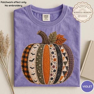 Op de afbeelding: Een violet t-shirt met een centrale pompoengrafiek bestaande uit diverse stofachtige patches met patronen zoals vleermuizen, manen, sterren, bloemen, pompoenen, spinnenwebben en ruiten. De tekst luidt: Patchwork effect only. No embroidery.