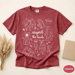 May include: Crimson-colored t-shirt with a white graphic featuring various cryptids and the text "respect the locals." The design includes creatures like Bigfoot, a Loch Ness Monster, and an alien.