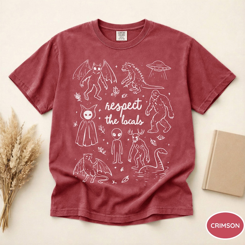 May include: Crimson-colored t-shirt with a white graphic featuring various cryptids and the text "respect the locals." The design includes creatures like Bigfoot, a Loch Ness Monster, and an alien.
