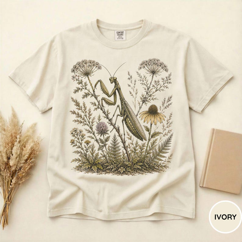 May include: Ivory-colored t-shirt featuring a detailed illustration of a praying mantis amidst wildflowers and foliage. The design includes a variety of plants and a yellow flower. The shirt has a classic crew neck and short sleeves. The word "IVORY" is printed on the bottom right.