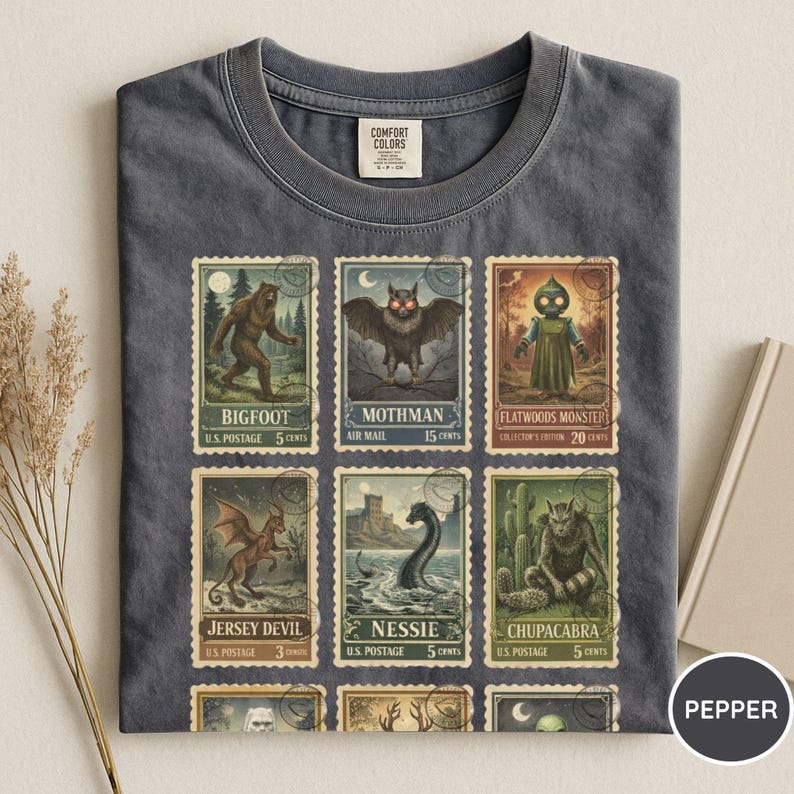 May include: Dark gray Comfort Colors t-shirt featuring a collection of vintage-style postage stamps. The stamps depict various cryptids, including Bigfoot, Mothman, and the Jersey Devil. The shirt has a Pepper color tag.