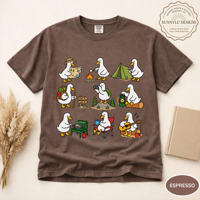 May include: A brown short-sleeve t-shirt featuring a graphic print of nine white ducks engaged in various camping activities, including hiking, roasting marshmallows, setting up a tent, using binoculars, and playing an acoustic guitar.