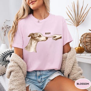 May include: A light pink t-shirt featuring a brown ink-style illustration of a dog's head being touched on the nose by a human finger. The text BOOP! is printed in brown above the dog's nose. The shirt has a crew neckline and short sleeves.