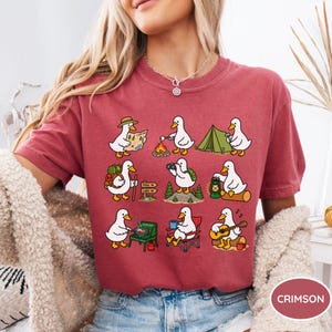 May include: A crimson t-shirt featuring a graphic print of nine white ducks engaged in various camping activities, including hiking, roasting marshmallows, setting up a tent, using binoculars, cooking on a stove, and playing an acoustic guitar.