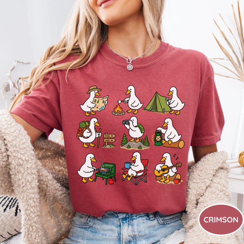 May include: A crimson t-shirt featuring a graphic print of nine white ducks engaged in various camping activities, including hiking, roasting marshmallows, setting up a tent, using binoculars, cooking on a stove, and playing an acoustic guitar.
