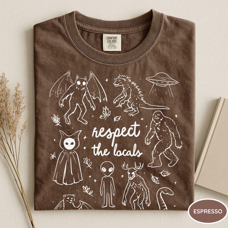 May include: Espresso-colored t-shirt with a crew neck. The shirt features a white graphic of various cryptids and a UFO, with the text "respect the locals". The shirt is made of a soft material.
