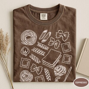 Puede incluir: Una camiseta de color marr&oacute;n oscuro con un gr&aacute;fico de l&iacute;neas blancas que muestra varias formas de pasta, incluyendo farfalle, ravioli, penne y l&aacute;minas de lasa&ntilde;a. La etiqueta interior del cuello muestra el texto Comfort Colors.