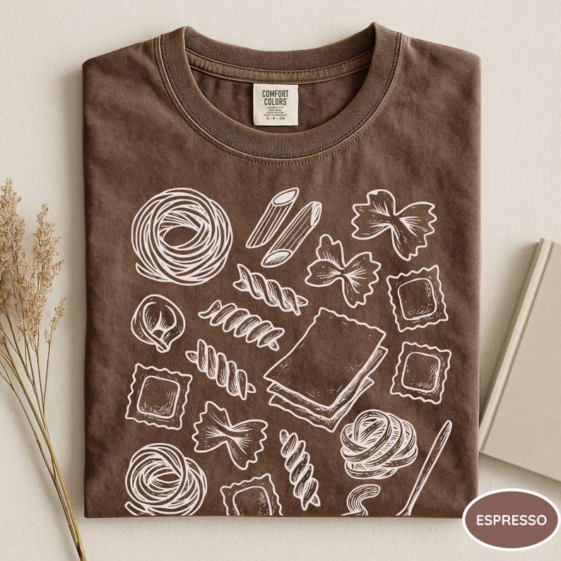 Puede incluir: Una camiseta de color marr&oacute;n oscuro con un gr&aacute;fico de l&iacute;neas blancas que muestra varias formas de pasta, incluyendo farfalle, ravioli, penne y l&aacute;minas de lasa&ntilde;a. La etiqueta interior del cuello muestra el texto Comfort Colors.