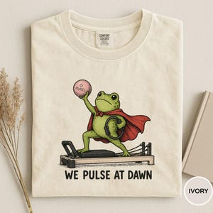 May include: Ivory-colored t-shirt featuring a cartoon frog in a red cape, holding a Pilates ball and a ring, standing on a Pilates reformer. The text "WE PULSE AT DAWN" is printed below the frog.