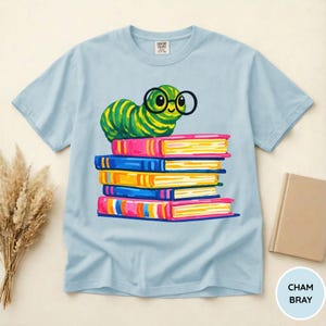 Pu&ograve; includere: Una t-shirt azzurro chiaro con la grafica di un bruco a strisce verdi e gialle che indossa occhiali tondi neri, appoggiato sopra una pila di cinque libri colorati. La maglietta presenta un girocollo e un'etichetta del marchio Comfort Colors.