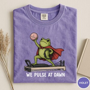 May include: A violet t-shirt featuring a graphic of a green frog wearing a red cape, holding a pink ball labeled "PILATES" and a black ring while standing on a reformer machine. The text "WE PULSE AT DAWN" is printed in white below the illustration.