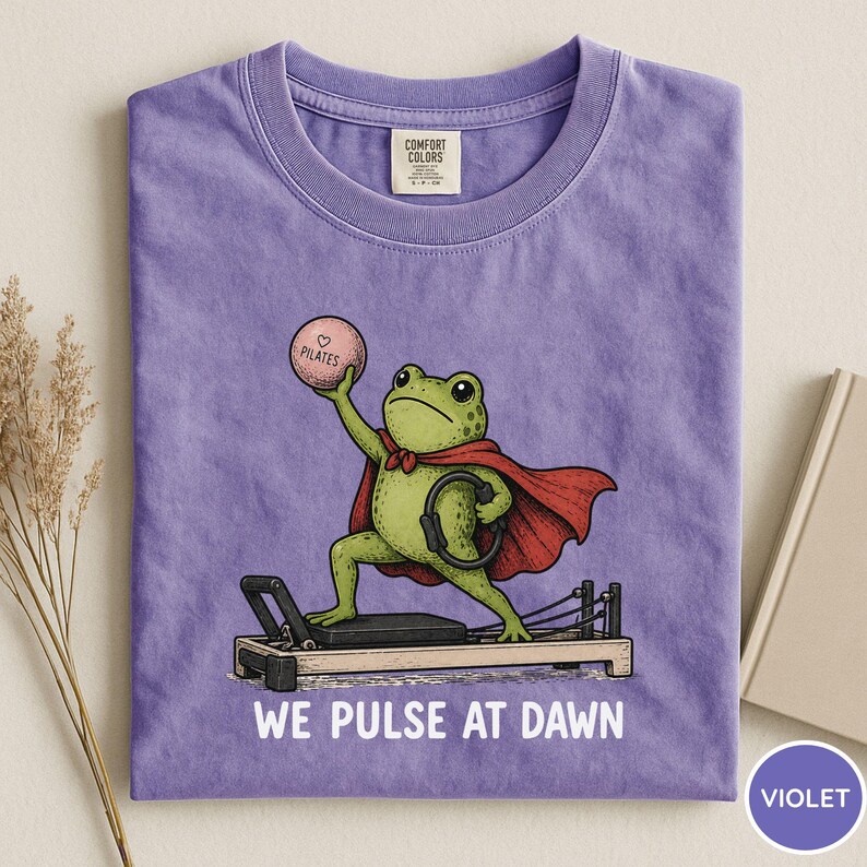 May include: A violet t-shirt featuring a graphic of a green frog wearing a red cape, holding a pink ball labeled "PILATES" and a black ring while standing on a reformer machine. The text "WE PULSE AT DAWN" is printed in white below the illustration.