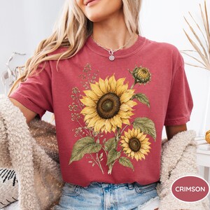 May include: Crimson-colored t-shirt featuring a detailed illustration of sunflowers with green leaves and a bud. The design includes a large, fully bloomed sunflower and a smaller one. The shirt is a short-sleeved crew neck.