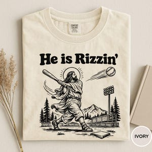 Könnte beinhalten: Elfenbeinfarbenes T-Shirt mit dem Text "He is Rizzin'" über einer Schwarz-Weiß-Illustration von Jesus beim Baseballspielen. Das Design umfasst einen Baseball, Stadionlichter und einen Berg im Hintergrund.