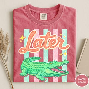 May include: A red t-shirt with a graphic of a green alligator and the word "Later" in orange script. The design includes vertical stripes in light blue and pink, with yellow star accents. The shirt is labeled "Watermelon".