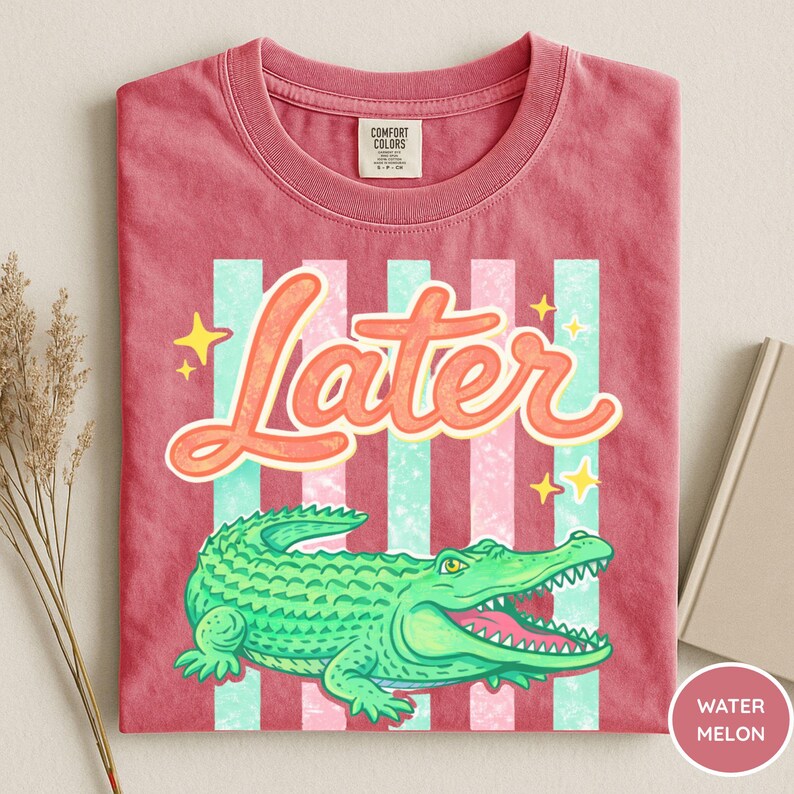 May include: A red t-shirt with a graphic of a green alligator and the word "Later" in orange script. The design includes vertical stripes in light blue and pink, with yellow star accents. The shirt is labeled "Watermelon".
