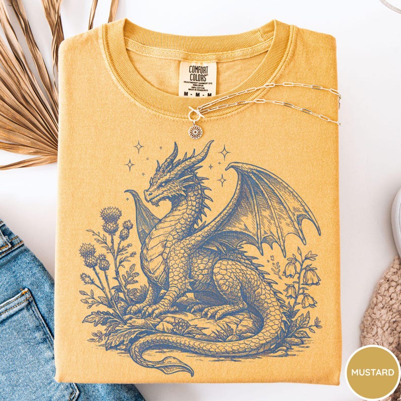 Peut inclure: T-shirt moutarde avec un motif de dragon bleu. Le dragon est entour&eacute; de fleurs et d'&eacute;toiles. Le t-shirt a un col rond et des manches courtes. Un collier dor&eacute; avec un pendentif est pos&eacute; sur le t-shirt.