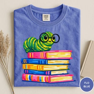 Pu&ograve; includere: Una t-shirt a maniche corte color blu pervinca con la grafica di un bruco verde che indossa occhiali tondi neri, appoggiato sopra una pila di cinque libri colorati. La maglietta presenta un'etichetta del marchio Comfort Colors sul collo.