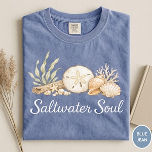 May include: Blue jean-colored t-shirt with a graphic of seashells, sand dollars, and coral. The words "Saltwater Soul" are printed in white script across the bottom. The shirt has a crew neck and a Comfort Colors tag.