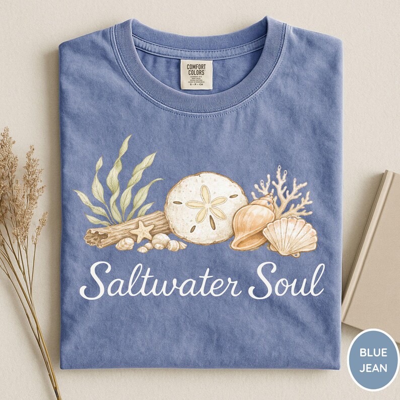May include: Blue jean-colored t-shirt with a graphic of seashells, sand dollars, and coral. The words "Saltwater Soul" are printed in white script across the bottom. The shirt has a crew neck and a Comfort Colors tag.