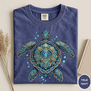 May include: A navy blue crew neck t-shirt featuring a central graphic of a sea turtle with a decorative shell pattern in shades of blue and gold. The turtle is surrounded by small blue bubbles. The inner neck label reads Comfort Colors.