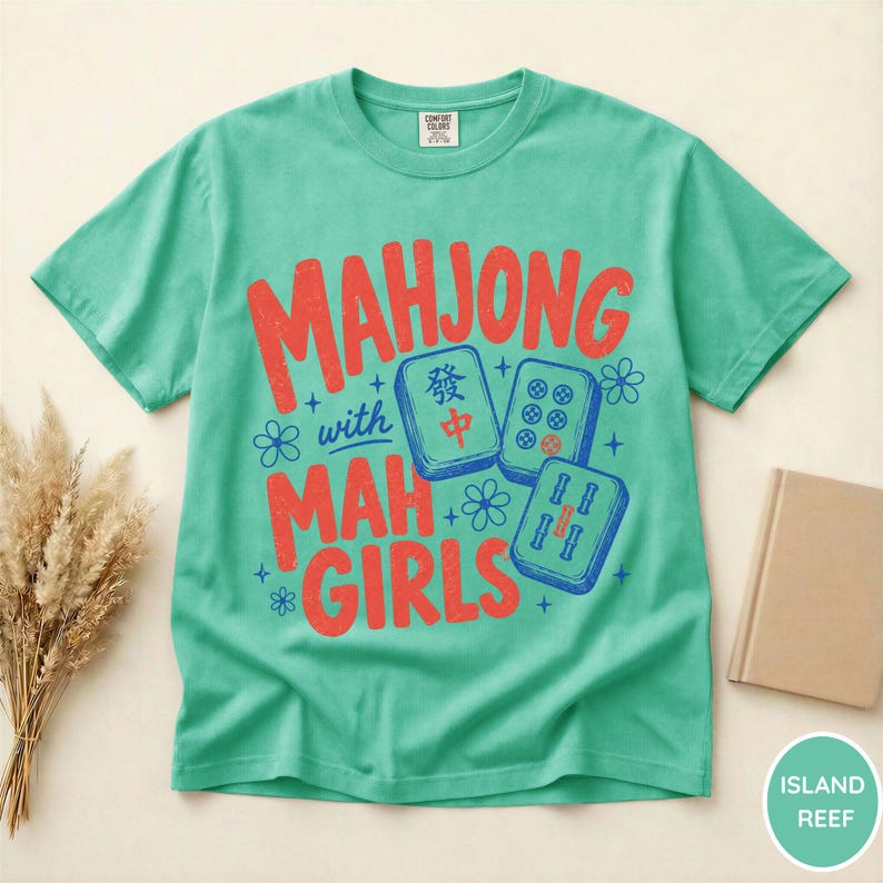 May include: A mint green short-sleeve t-shirt featuring the red text "MAHJONG with MAH GIRLS" and three blue mahjong tiles. The design includes small blue star and flower accents. The shirt has a crew neckline and a Comfort Colors brand label.