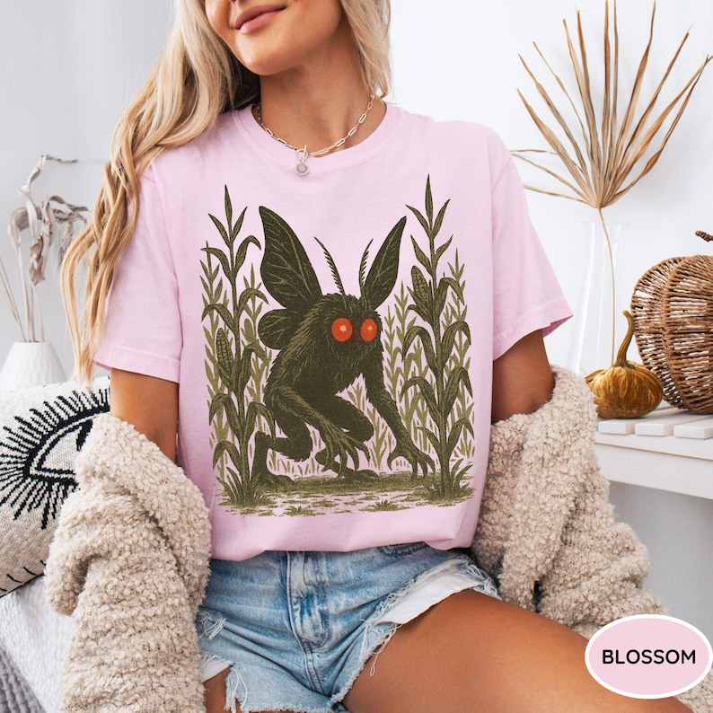K&ouml;nnte beinhalten: Hellrosa T-Shirt mit einem dunkelgr&uuml;nen Kreaturen-Design mit roten Augen und gro&szlig;en Fl&uuml;geln, umgeben von gr&uuml;nen Maisst&auml;ngeln. Das T-Shirt wird von einer Person getragen.