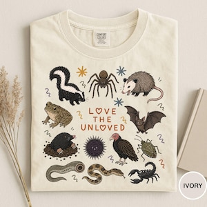 Puede incluir: Camiseta de color marfil con un diseño de varios animales y el texto "LOVE THE UNLOVED". El diseño incluye una mofeta, una araña, una zarigüeya, una rana, un topo, un erizo de mar, un murciélago, un escarabajo, un escorpión y una serpiente. Cuello redondo y mangas cortas.