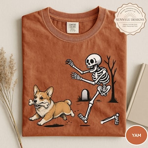 May include: A rust-colored t-shirt featuring a graphic of a corgi running with a bone in its mouth while being chased by a skeleton near a tombstone and a bare tree. The shirt has a crew neck and a Comfort Colors brand label at the inner collar.