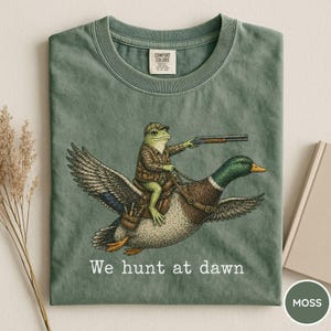 Puede incluir: Camiseta verde musgo con una ilustraci&oacute;n humor&iacute;stica de una rana con atuendo de caza montada en un pato y sosteniendo una escopeta. El texto "We hunt at dawn" est&aacute; impreso debajo. Cuello redondo y mangas cortas.