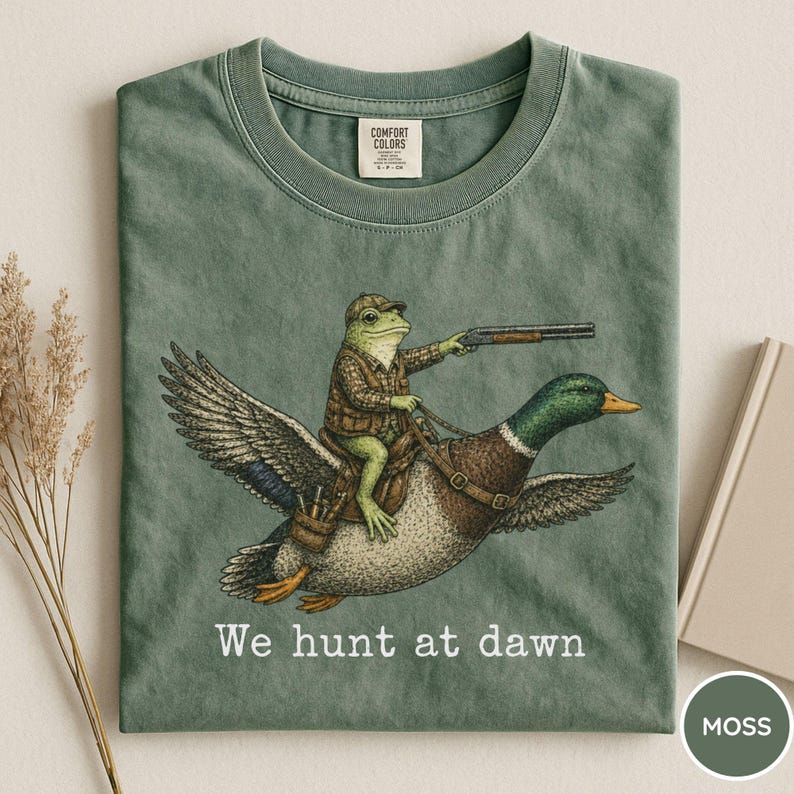 Puede incluir: Camiseta verde musgo con una ilustraci&oacute;n humor&iacute;stica de una rana con atuendo de caza montada en un pato y sosteniendo una escopeta. El texto "We hunt at dawn" est&aacute; impreso debajo. Cuello redondo y mangas cortas.