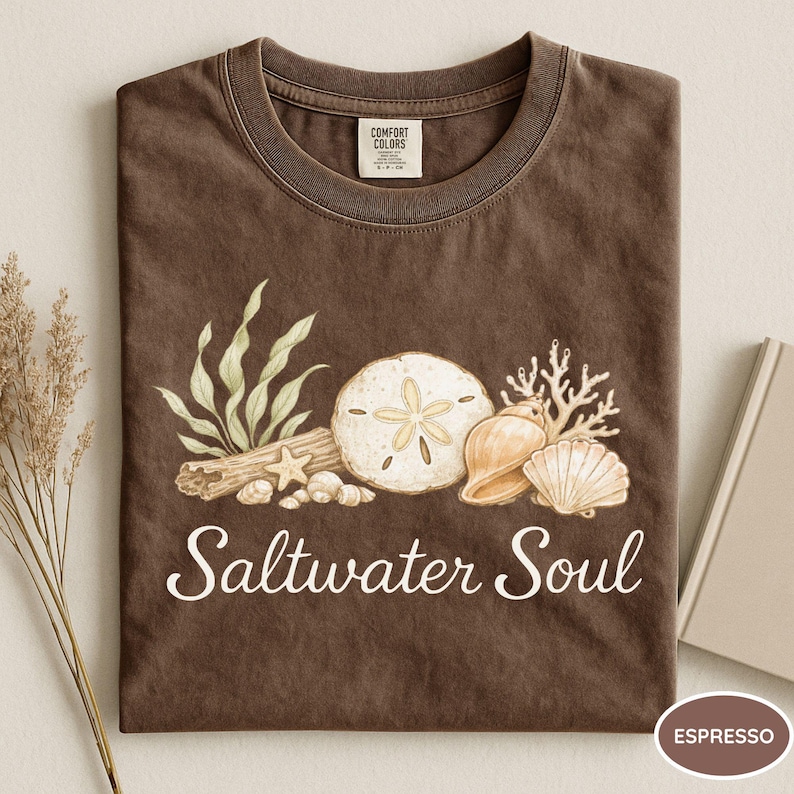 May include: Espresso-colored t-shirt with a round neckline. The shirt features a watercolor design of seashells, sand dollars, and seaweed, with the words "Saltwater Soul" in a script font. A book and dried plant are in the background.