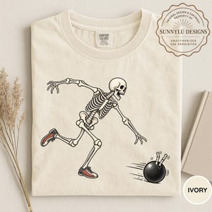 Op de afbeelding: Een ivoorkleurig t-shirt met een grafische illustratie van een skelet in een dynamische bowlinghouding. Het skelet draagt rood-witte bowlingschoenen en werpt een zwarte bowlingbal met bewegingslijnen die erachteraan lopen.
