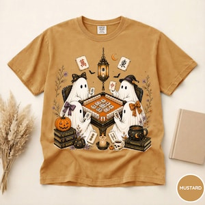 May include: A mustard yellow t-shirt featuring a graphic of four ghosts playing mahjong around a table. The design includes a hanging lantern, books, a pumpkin, a skull mug, and mahjong tiles with Chinese characters. The shirt has a crew neck.