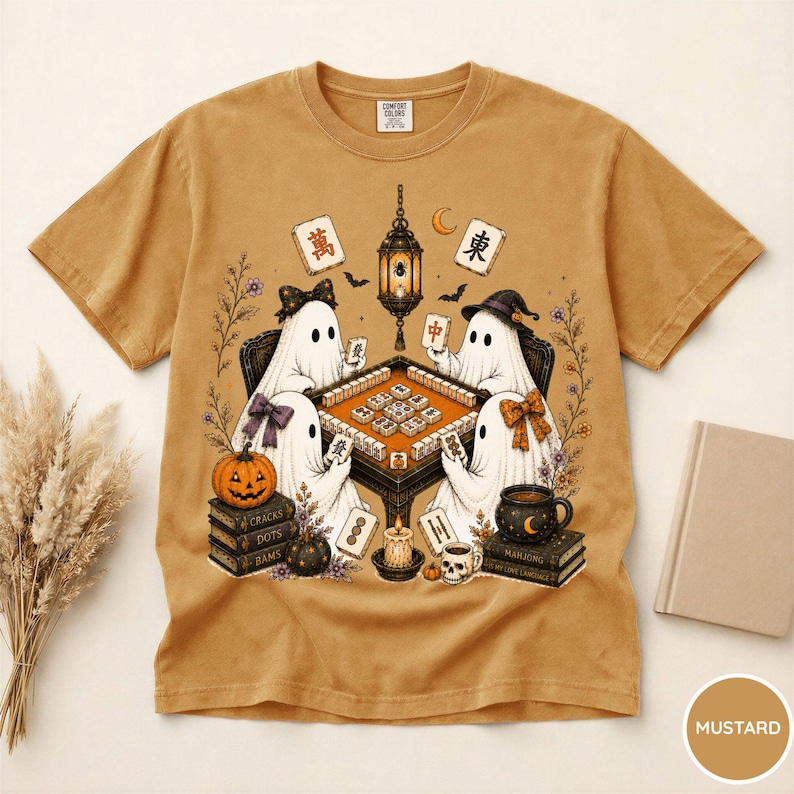 May include: A mustard yellow t-shirt featuring a graphic of four ghosts playing mahjong around a table. The design includes a hanging lantern, books, a pumpkin, a skull mug, and mahjong tiles with Chinese characters. The shirt has a crew neck.
