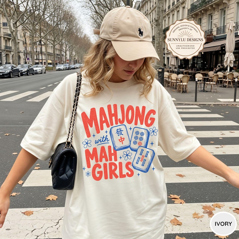 May include: An ivory t-shirt featuring red text that reads "MAHJONG with MAH GIRLS" alongside blue illustrations of mahjong tiles and small flower accents. The shirt is paired with a black quilted shoulder bag and a beige baseball cap.
