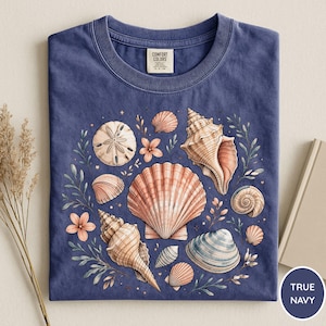 May include: A navy blue t-shirt featuring a seashell design. The design includes various seashells in shades of peach, tan, and blue, surrounded by green foliage and small pink flowers. The shirt has a classic crew neck and a tag that says "Comfort Colors".