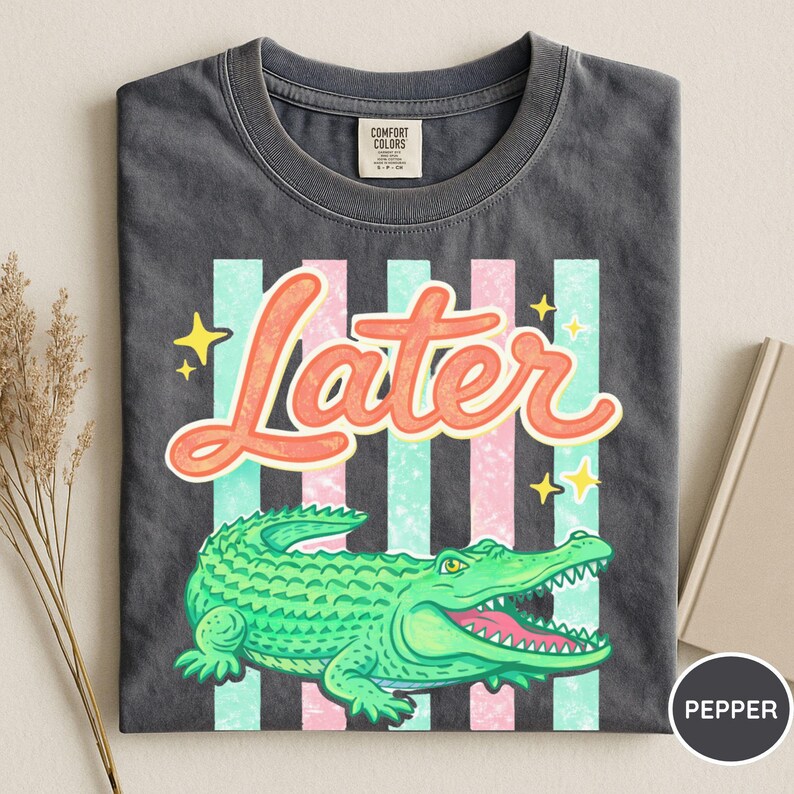 May include: Dark gray t-shirt with a graphic of a green alligator, the word "Later" in orange script, and vertical stripes in pink and teal. The shirt has a crew neck and a Comfort Colors tag.