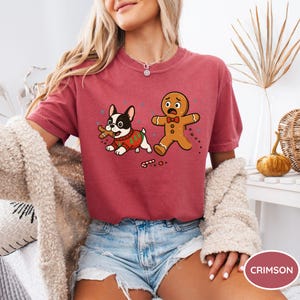 May include: Crimson-colored t-shirt featuring a cartoon dog in a Christmas sweater chasing a gingerbread man. The dog is black and white, and the gingerbread man has a worried expression. The shirt is paired with denim shorts.