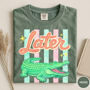 May include: Moss green t-shirt with a graphic of a green alligator and the word "Later" in orange script. The design includes pink and teal stripes and yellow stars. The shirt is folded.
