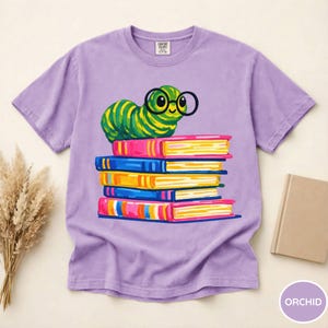 Pu&ograve; includere: Una t-shirt a maniche corte color viola chiaro con la grafica di un bruco a strisce verdi e gialle che indossa occhiali tondi neri, appoggiato sopra una pila di cinque libri colorati nelle tonalit&agrave; del rosa, blu e giallo.