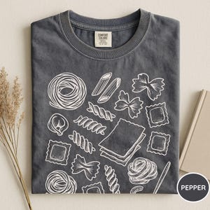 Puede incluir: Una camiseta de cuello redondo en color gris carb&oacute;n con un estampado gr&aacute;fico de l&iacute;neas blancas que muestra varias formas de pasta, incluyendo penne, farfalle, raviolis y l&aacute;minas de lasa&ntilde;a. Incluye la etiqueta de la marca Comfort Colors en el cuello.