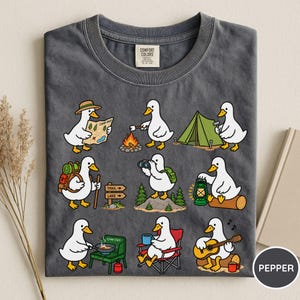 May include: A charcoal gray t-shirt featuring a graphic print of nine white ducks engaged in various camping activities, including reading a map, roasting marshmallows, setting up a tent, hiking, using binoculars, holding a lantern, cooking, sitting in a chair, and playing guitar.