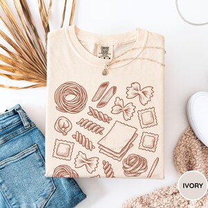 Puede incluir: Una camiseta de color crema con un gr&aacute;fico de l&iacute;neas marrones de varias formas de pasta, incluyendo farfalle, penne, ravioli y l&aacute;minas de lasa&ntilde;a. La camiseta tiene un cuello redondo y una etiqueta de la marca Comfort Colors en el interior.