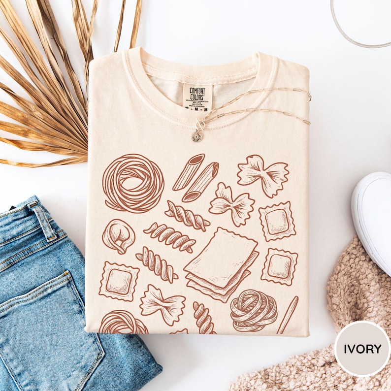 Puede incluir: Una camiseta de color crema con un gr&aacute;fico de l&iacute;neas marrones de varias formas de pasta, incluyendo farfalle, penne, ravioli y l&aacute;minas de lasa&ntilde;a. La camiseta tiene un cuello redondo y una etiqueta de la marca Comfort Colors en el interior.