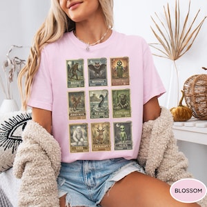 May include: A light pink t-shirt featuring a grid of cryptid illustrations, including Bigfoot, Mothman, and the Jersey Devil. Each image is framed with a vintage postage stamp design. The shirt is a casual, short-sleeved style.