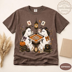 May include: An espresso-colored t-shirt featuring a graphic illustration of four white ghosts playing a mahjong game at a table. The design includes a hanging lantern, a pumpkin, stacks of books, a skull mug, and various mahjong tiles.