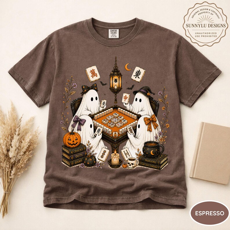 May include: An espresso-colored t-shirt featuring a graphic illustration of four white ghosts playing a mahjong game at a table. The design includes a hanging lantern, a pumpkin, stacks of books, a skull mug, and various mahjong tiles.