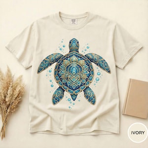 May include: An ivory short-sleeve t-shirt featuring a central graphic of a sea turtle with an intricate blue and gold mandala-style shell pattern, surrounded by small blue bubbles. The shirt has a crew neckline and a Comfort Colors brand label.